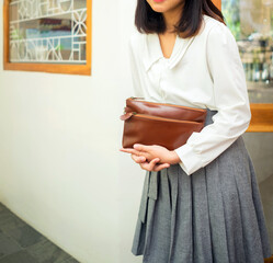The body of a girl in a gray skirt and white shirt stands on the side of the glass window while holding an elegant tan leather handbag.
