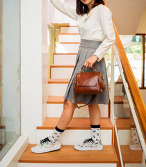 The body of a girl in a gray skirt and white shirt is standing in front of the stairs with a beautiful dark brown leather bag in her hand.