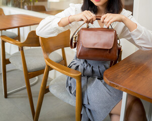 The body of a girl in a gray skirt and white shirt sitting in a cafe while showing off a beautiful dark brown leather bag