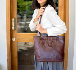 The body of a girl in a gray skirt and white shirt carrying a dark brown leather bag, standing in front of a glass window.