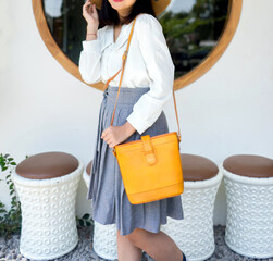 The body of a girl in a gray skirt and white shirt carrying a turmeric yellow leather bag, standing in front of a glass window.