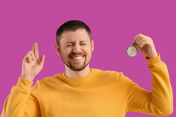 Young man with stopwatch crossing fingers on violet background