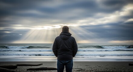 A man standing on a beach, gazing out at the ocean with a cloudy sky above.