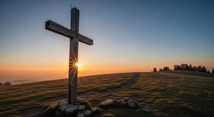 A wooden cross stands on a grassy hilltop at sunset, with the sun setting behind it.