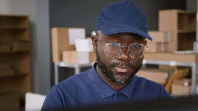 Medium close-up of face of busy African American male customer service associate working on computer at courier delivery company office, typing and analyzing shipment information on screen