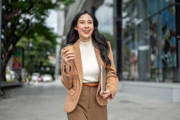 Pretty asian woman office worker holding coffee cup carrying laptop as walking outside shopping mall