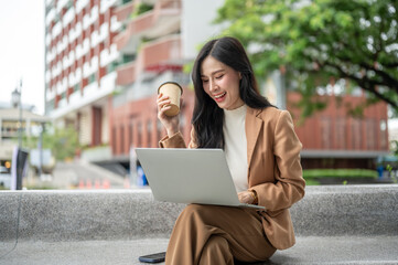 Pretty asian woman office worker holding coffee looking at laptop sitting on stone bench in a garden