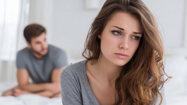 A woman sits on a bed looking pensive, while a man in the background appears troubled. The soft morning light highlights the tension in their relationship.