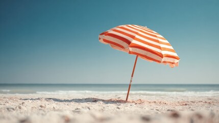 Striped umbrella rests on the soft sand of a peaceful beach, with the calm ocean waves gently lapping at the shore under a clear blue sky on a sunny day.