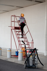 Maintenance worker in uniform and helmet climbing a red mobile ladder stand near cleaning equipment.