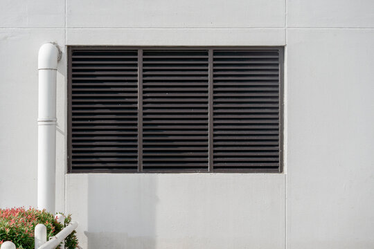 Black metal louvered ventilation grille on a white wall with red photinia leaves in the corner.