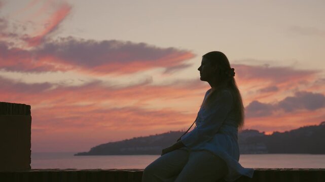 Wide side profile silhouette of a woman seated calmly on a terrace ledge, framed by dramatic pink and orange clouds. Stillness and quiet awe - Powered by Adobe