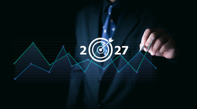 Businessman pointing at digital target icon with 2027 and rising line graph, symbolizing future strategy, financial growth, data analytics, corporate planning, and market performance trends.
