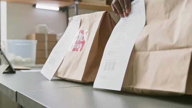 Close-up of hands of anonymous African American employee placing packed bag with stapled invoice, containing grocery order, on counter at dark store or online supermarket, ready for delivery or pickup