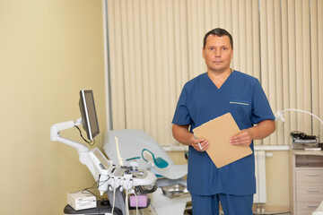 Horizontal photo. Man doctor in blue uniform stands with paper with patient information and looks at the camera in gynecology hospital office with ultrasound machine in private clinic. Health care