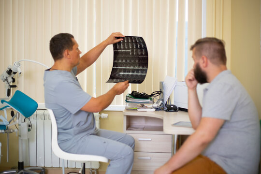 Horizontal photo. Man doctor in blue uniform gives diagnosis, shows radiology x-ray shot to man with beard in hospital office of clinic. Concept of medicine aid, treatment, work, health care, illness