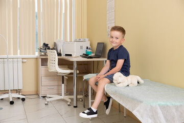 Horizontal photo. Five year old boy sits on couch during pediatrician doctor appointment, smiles, looks at camera. Children in hospital clinic office. Concept of health care, medicine, heal, work, aid