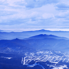 Panoramic winter view of Sheregesh ski resort in Russia see from snowy mountain Mystag slope. Snow-covered forest, village and hotels, distant peaks on horizon under cloud blue sky, tonal perspective.