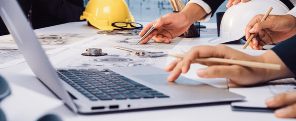 A group of engineers engages in a detailed planning session around blueprints and technical drawings, using laptops, tools, and safety gear, representing teamwork and innovation. SACTR