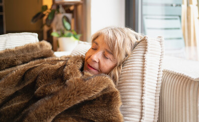 Senior woman sleeping peacefully on sofa, covered by a soft brown faux fur blanket