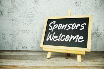 wooden easel displays a blackboard with the bold white text "Sponsors Welcome" set against a rustic wooden surface and textured gray wall background