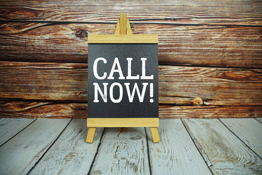 wooden easel holds a blackboard with the white text "Call Now" set against a rustic wooden wall and light-colored wooden floor