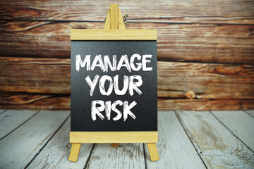 wooden easel holds a blackboard with the white text "Manage Your Risk" set against a rustic wooden wall and light-colored wooden floor