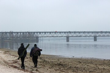 Two fishermen with rods walk along a sandy riverbank near a steel bridge on a foggy morning in Kremenchuk.