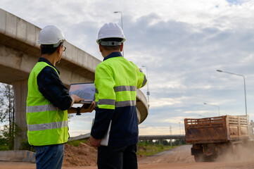 Civil engineers reviewing plans on laptop and supervising road construction while monitoring dump truck movement at infrastructure site.