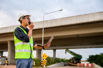 Civil engineer communicating with walkie talkie while supervising road construction at elevated highway site. Inspect project, supervise new infrastructure work in Thailand Asia.