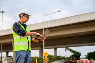 Construction engineer using laptop to supervise project at elevated road construction site. Inspect road construction, civil engineer working outdoors in Thailand Asia.