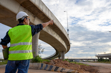 Construction engineer supervising elevated road construction, inspecting structure and directing work at infrastructure site with walkie talkie and safety gear.