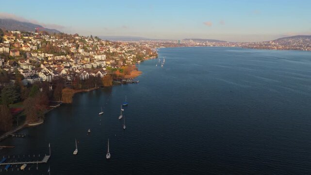 Drone glides over deep blue Lake Zurich toward Zurich. View of lakeside houses, sailboats, and distant Zurich skyline on a bright, calm day.