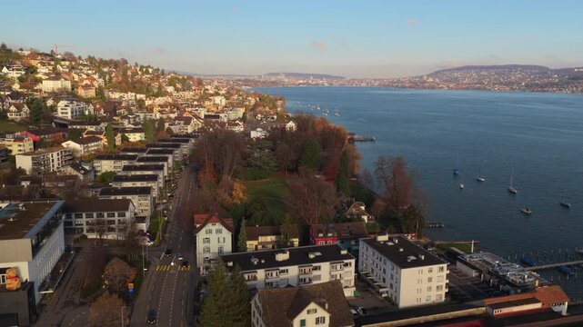 Drone footage showing Kilchberg and the Lake Zurich shoreline on a clear calm day. The camera reveals urban architecture, parks, and boats along the lake. Ideal for travel and cityscape projects.