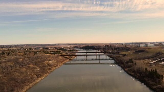 North Saskatchewan River flows toward Edmonton as soft daylight reveals distant bridges, open prairie slopes, and calm water with light ice patches creating wide peaceful scene from steady drone view.