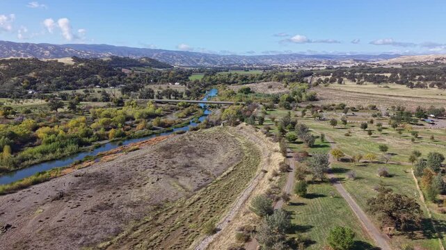 Drone performs a wide panning move above dirt roads, dry mounds and Cache Creek, revealing distant hills under a clear sky in Capay, California.