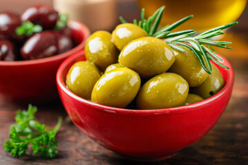 Glossy green olives garnished with fresh rosemary in a vibrant red bowl with blurred background featuring dark olives and fresh herbs on rustic wooden surface