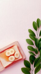 A slice of toast topped with honey and banana slices sits on a wooden board, next to a sprig of green leaves against a pink background.