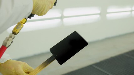 Professional auto body technician applies black paint onto a small test panel using a spray gun inside a clean, well lit automotive paint booth. - Powered by Adobe