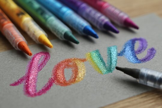 Drawing colorful love text using crayons to celebrate LGBTQ pride month on a wooden surface with vibrant rainbow hues