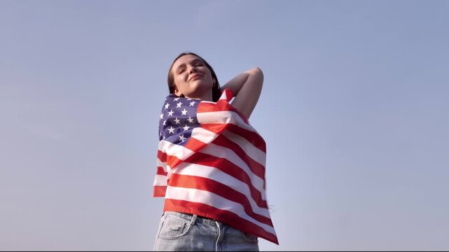A young woman holds and waves a large American flag against a clear blue sky. Her hair blows in the wind, symbolizing freedom, patriotism, and pride in the USA.
