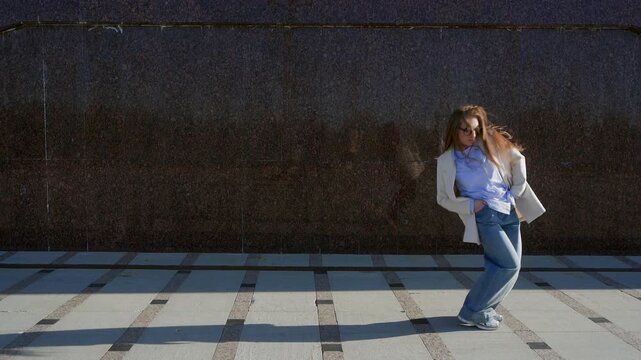 Energetic female in relaxed jeans and blazer dancing contemporary modern dace walks purposefully outdoors, sunlight casting defined shadows on granite wall, emphasizing motion.