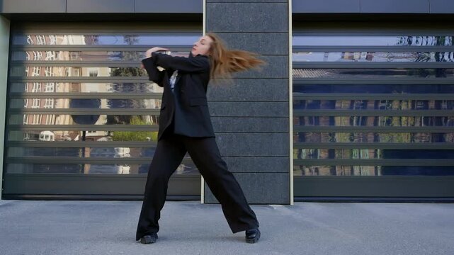 Confident female in elegant black suit and black pants dancing contemporary modern dace against gray striped building facade, showing poise and modern style outdoors.