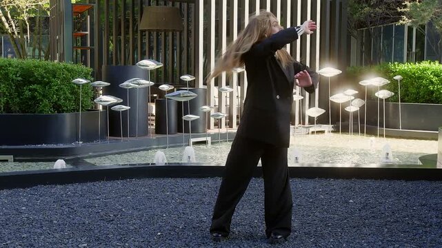 Dynamic dancer in black suit performs creative movement near modern fountain with metal sculptures and greenery in urban courtyard dancing contemporary modern dace