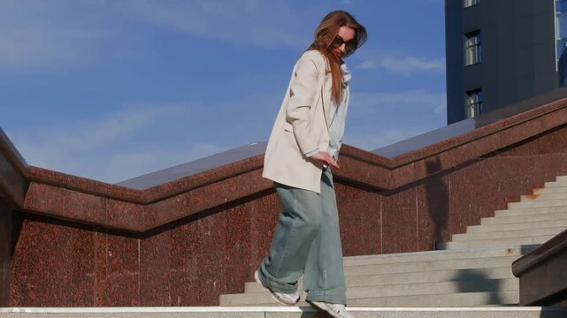 Confident woman dancing contemporary modern dace in oversized casual jeans, white sneakers, blazer, and blue shirt stands on modern city stairs, sunlight highlighting urban mood.