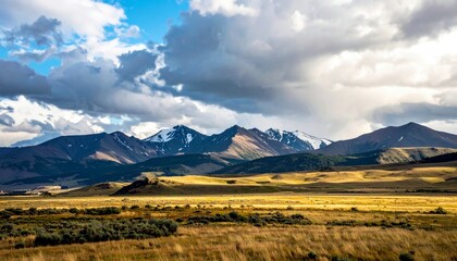 A vast, sunlit valley with golden grass leads to a range of rugged mountains, some capped with snow, under a dramatic, cloudy sky.