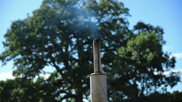 Farm tractor exhaust pipe releasing light smoke under clear blue sky and green trees. Engine emissions and mild air pollution often associated with older farm equipment. 