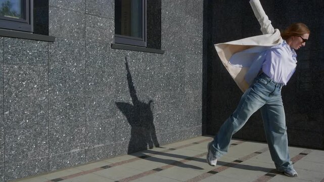 Dynamic dancing woman dancing contemporary modern dace leans back in loose jeans, white sneakers, light blazer and blue shirt, conveying tension and creativity outside a stone building.