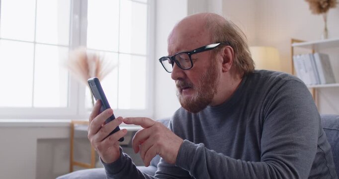 Elderly man with glasses sitting on sofa and staring at smartphone with confused and frustrated expression. Senior male struggling to understand how to use mobile application or internet at home.
