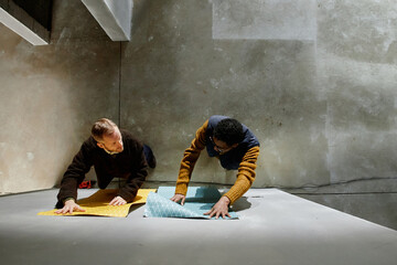 Caucasian middle aged man and Black young adult man arranging patterned paper sheets on concrete wall, both focused on collaborative creative project in industrial setting